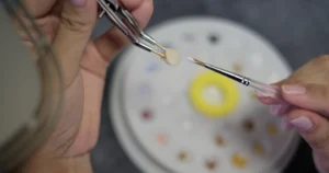 Dental technician holding a CEREC® crown with tweezers while applying glaze or color using a small brush, with a shade palette in the background in Las Vegas, NV.